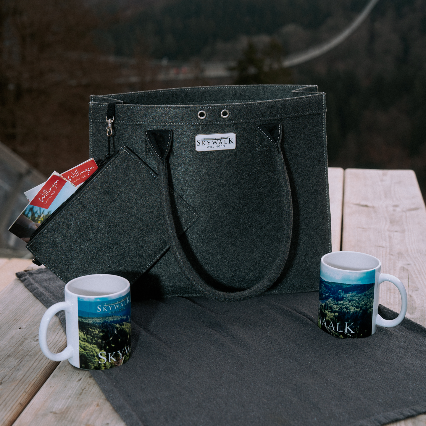 Bild: Es zeigt eine Produktkombination aus einer Filztasche und zwei Tassen. Sie stehen auf einem Holztisch in der Natur. In dem Etui, der kleinen Zusatztasche schauen die Spitzen von Willinger Wanderkarten heraus.