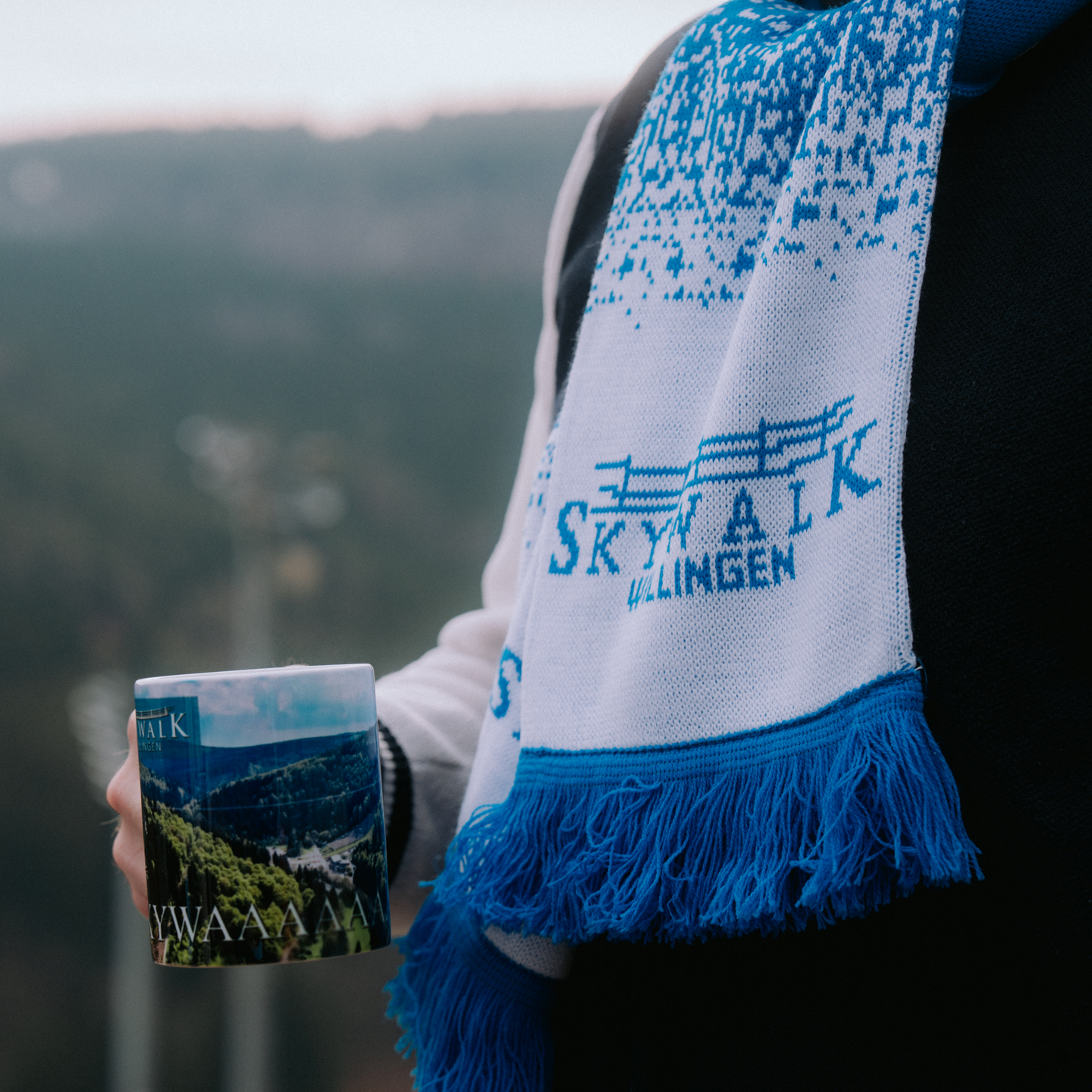 Es zeigt den Schal an einer Person umgebunden. Die Person hält ebenfalls eine Skywalk-Willingen Tasse mit Luftbild in der Hand.