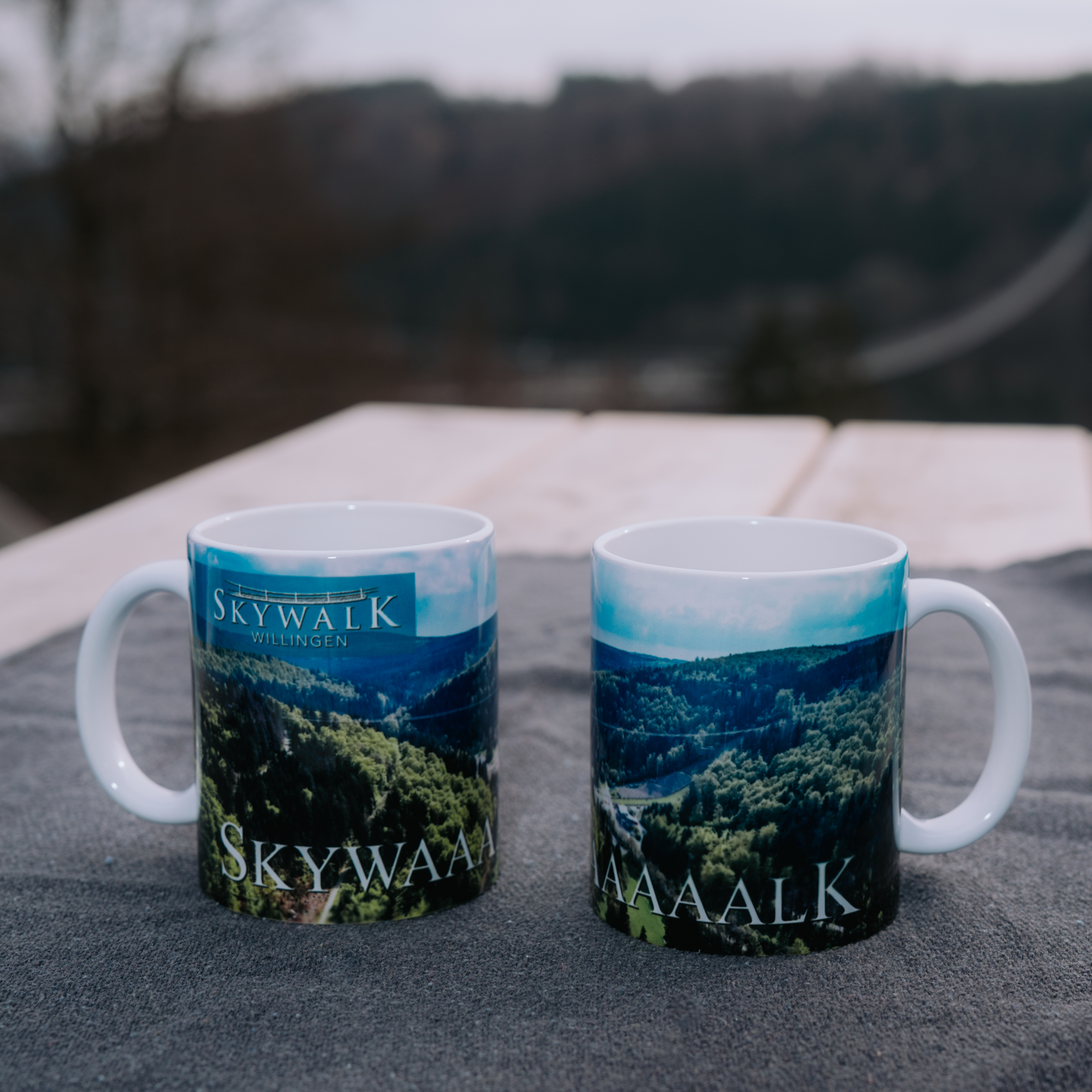 Bild: Es zeigt zwei weiße Bildmotivtassen vom Skywalk-Willingen mit Henkel links und Henkel rechts auf einem Tisch in der Natur. Die Tasse ist jeweils von innen sowie der Henkel der Tasse sind weiß. Außen ist die Tasse vom Luftbild umschlossen.